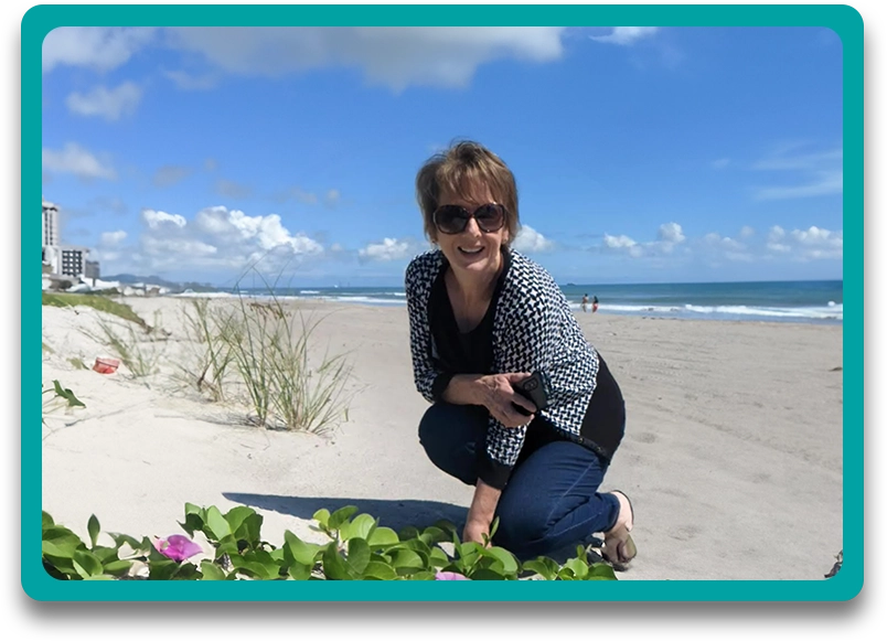 A woman kneeling down on the beach next to flowers.